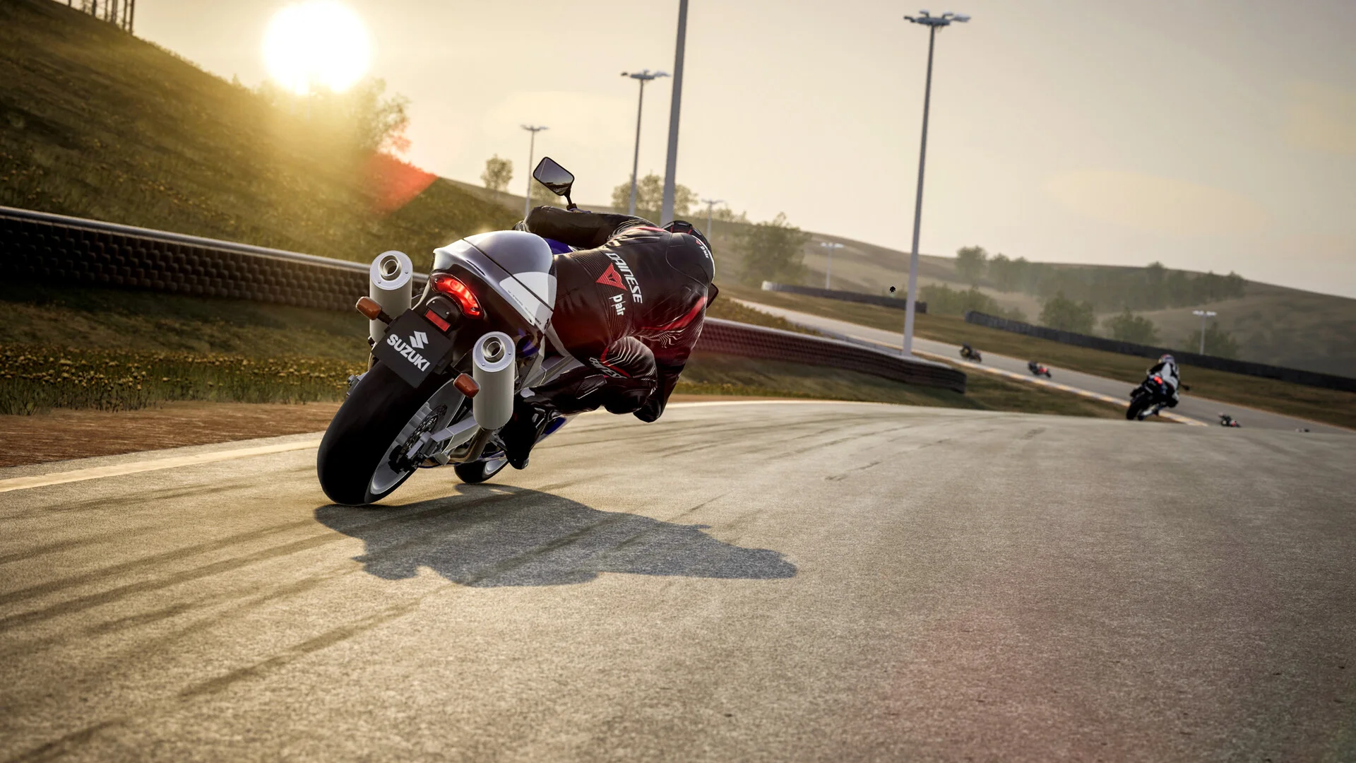 White-and-blue Suzuki superbike from behind, rider in a black Dainese suit carving a sunlit hill circuit with other bikes ahead