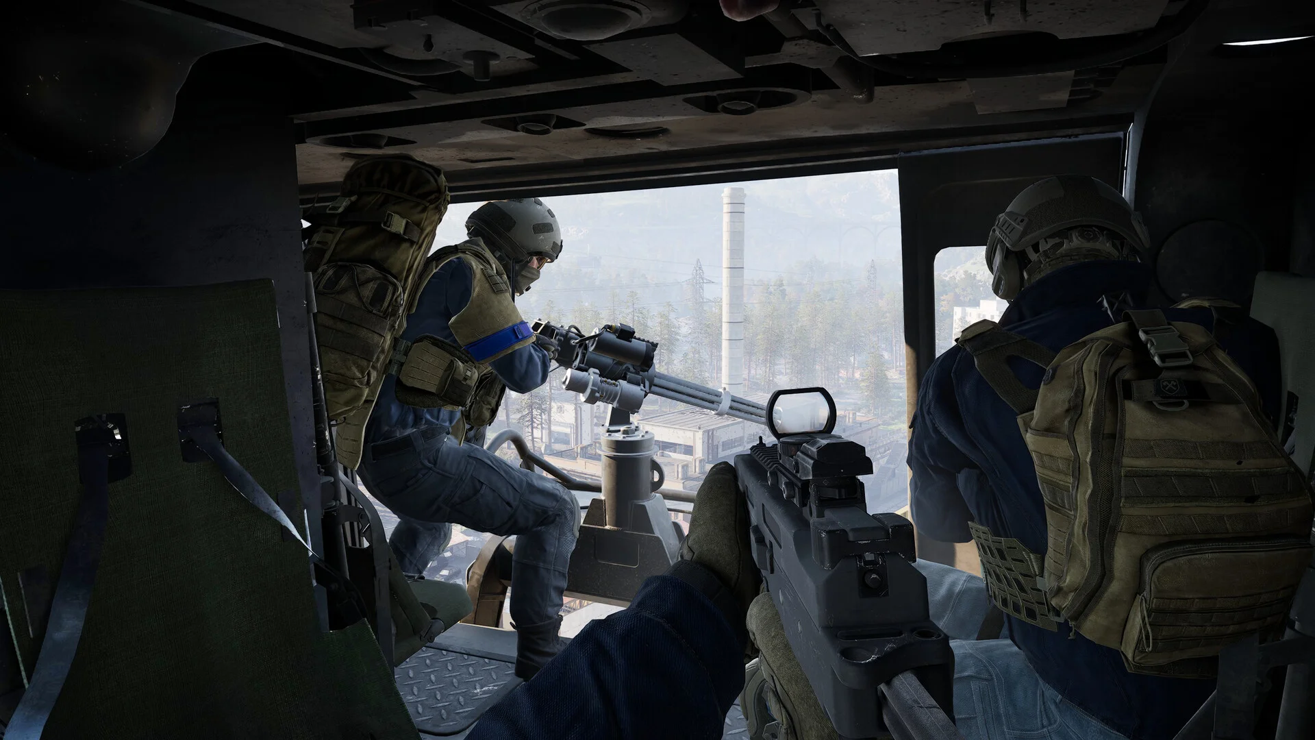 First-person in a helicopter cabin: door gunner on the left, rifle with red-dot sight in the foreground, factory chimney ahead