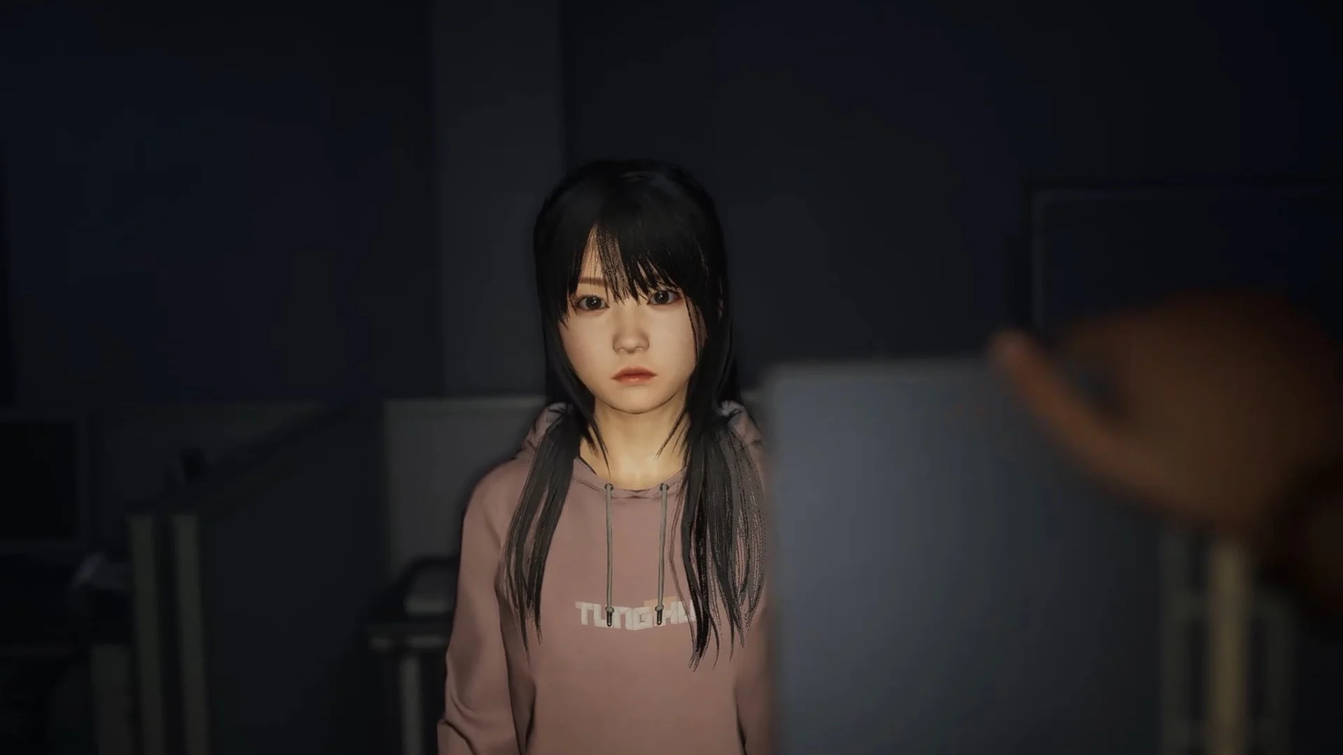 Student in a pink hoodie standing in a dark office looking at the player
