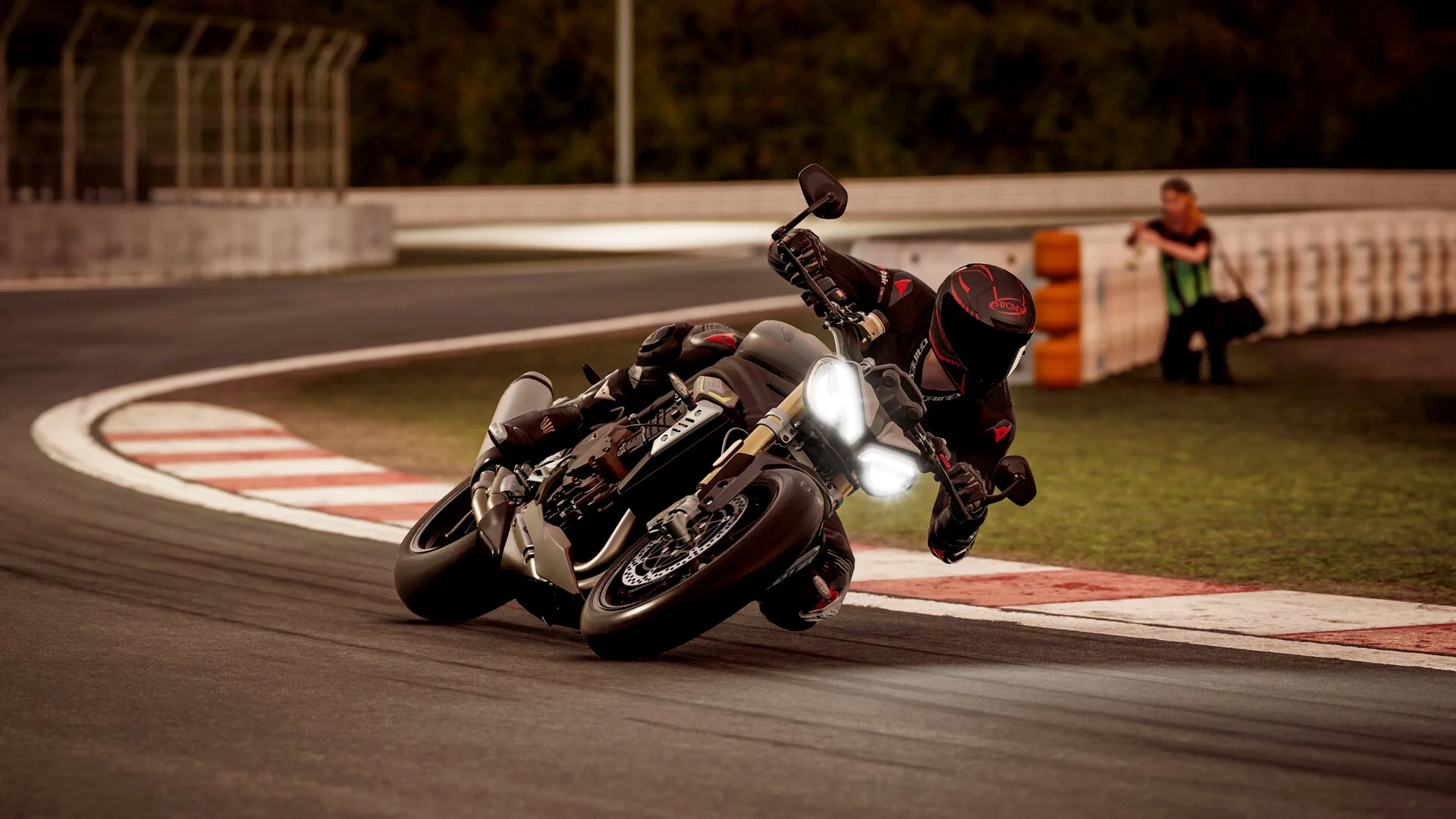 Grey naked bike leaned hard on a red-and-white kerb, rider’s knee out, a marshal with a camera beyond the barrier