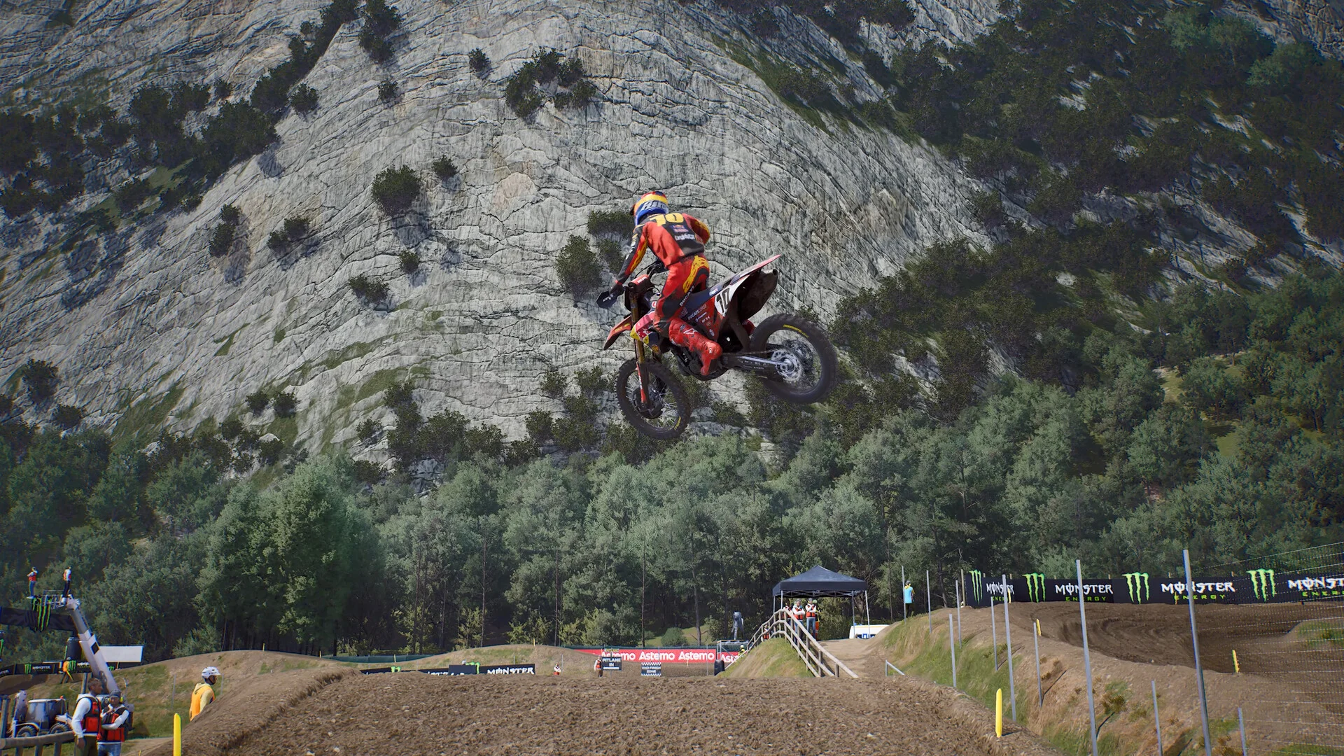 Orange-and-red rider number 93 jumps a dirt table against a cliff face, Monster Energy banners along the track