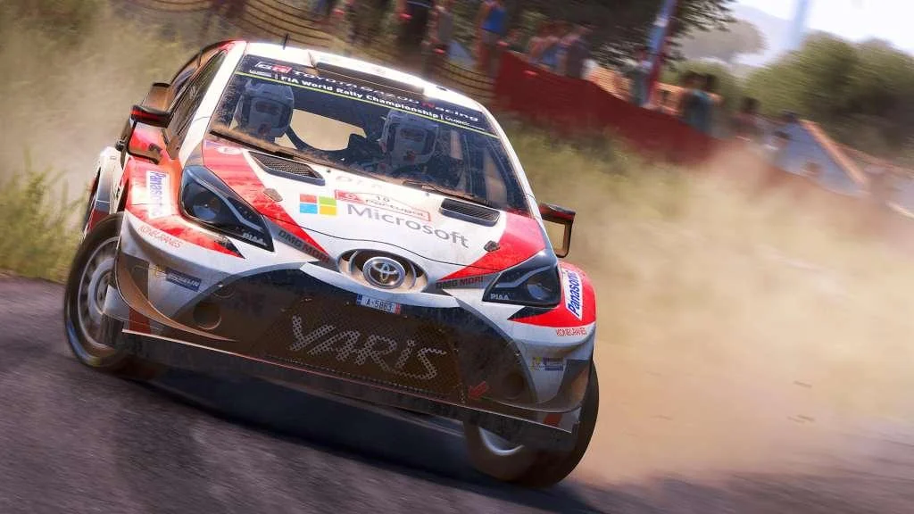 White-red Toyota Yaris rally car with roof lights and two helmeted crew, sliding on dirt past spectators and a red barrier