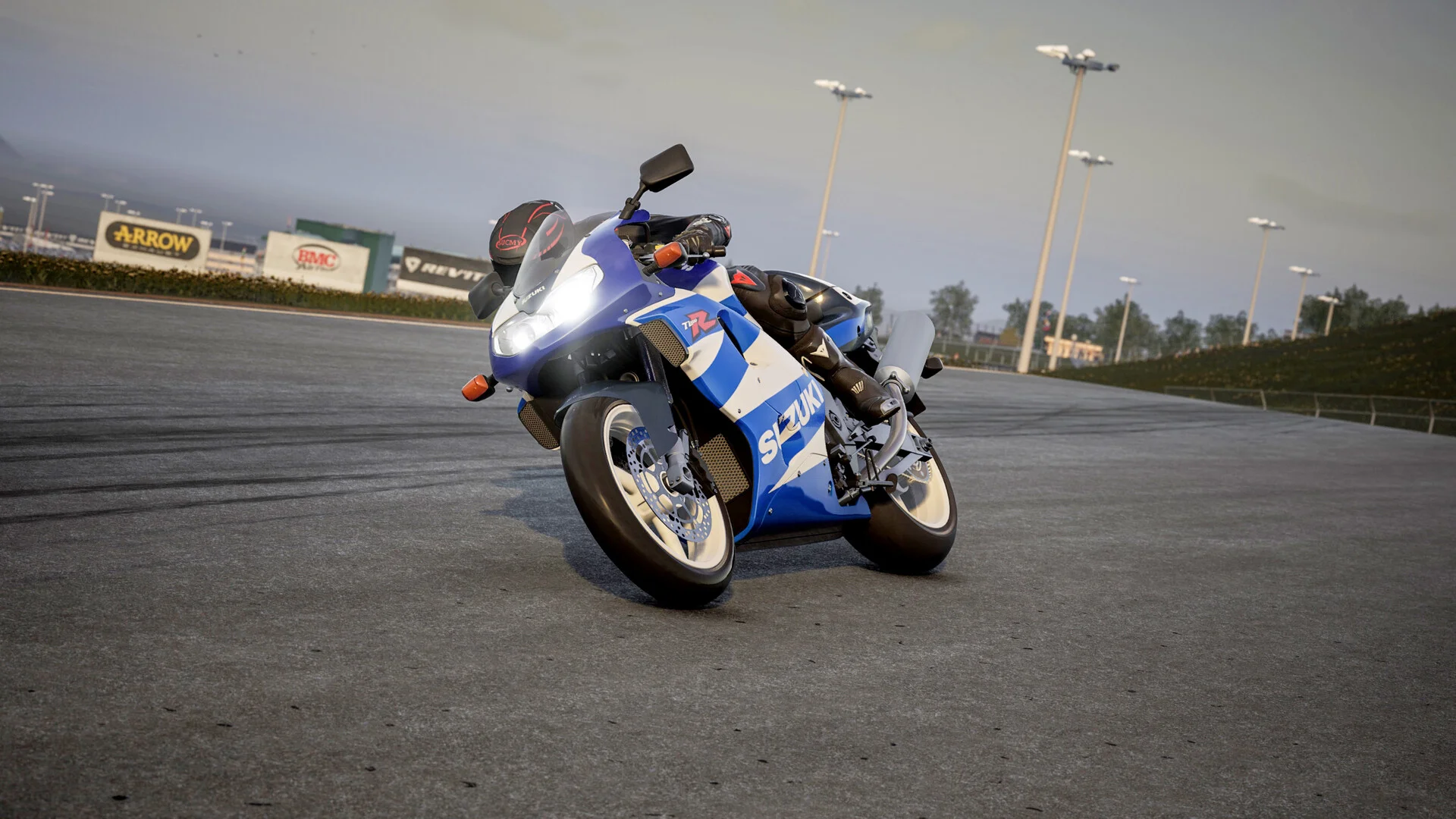Blue-and-white Suzuki 7L posed on empty tarmac, rider still aboard, Arrow and BMC hoardings under a pale sky