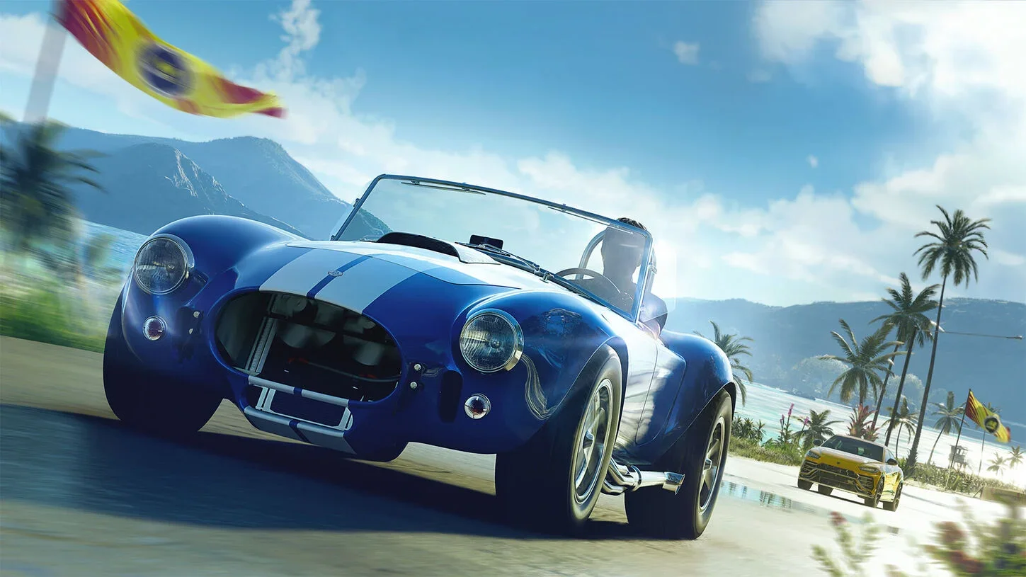 Blue-and-white striped open-top Cobra convertible leads a yellow supercar along a palm-lined beach road with mountains behind